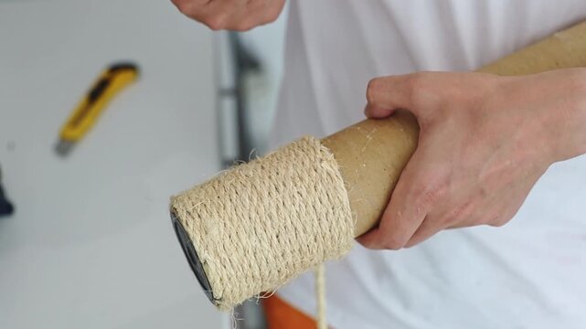 One young unrecognizable guy, while repairing, glues a new jute thread onto a cardboard tube of a cat's house, pressing it with his fingers, and, getting burned, pulls it away, standing at an angle in