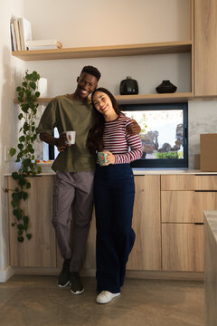Diverse couple standing in modern kitchen holding ceramic mugs, leaning, wearing casual clothes