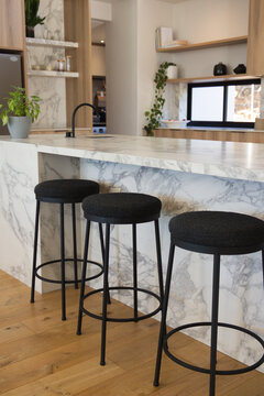 Three black bar stools are lining marble waterfall island, matte black faucet, wood shelves