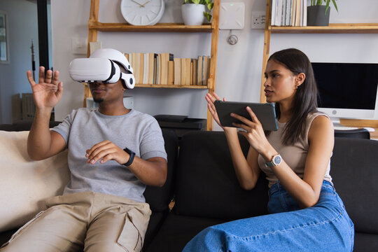African American couple wearing white VR headset, using tablet, sitting on dark sofa in living room