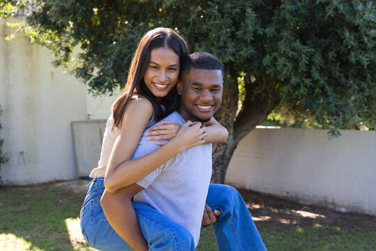 African American couple carrying woman piggyback and smiling in yard with tree, wall, sunlit grass
