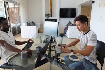 Obraz premium Diverse male coworkers working at glass table in living room using laptops, monitor, headphones