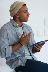 African American man sitting on white seat at home wearing tan beanie holding tablet gesturing © WavebreakMediaMicro