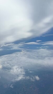 Aerial view of fluffy white clouds and blue sky captured from an airplane window during flight over a landscape with visible land. Slow motion vertical footage, 4K, copyspace