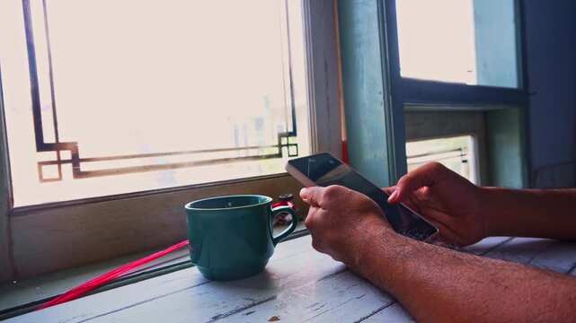 A person sits comfortably at a sunlit window desk holding a modern mobile phone while enjoying a warm cup of coffee during a peaceful moment of digital connection and morning relaxation at home