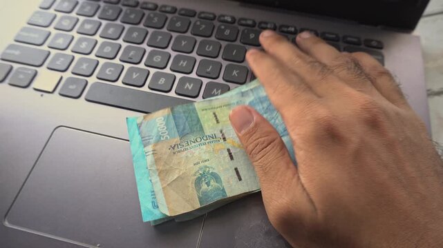 Close up of hand holding a stack of Indonesian rupiah banknotes over laptop keyboard, symbolizing online business, earning profit, internet income, digital banking and e-commerce transactions.