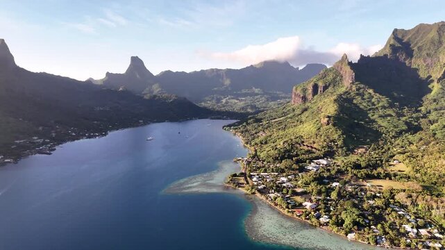 Aerial drone shot pulling back over a deep blue bay, revealing the coastal village, turquoise lagoon reef flats, and majestic volcanic peaks of Moorea
