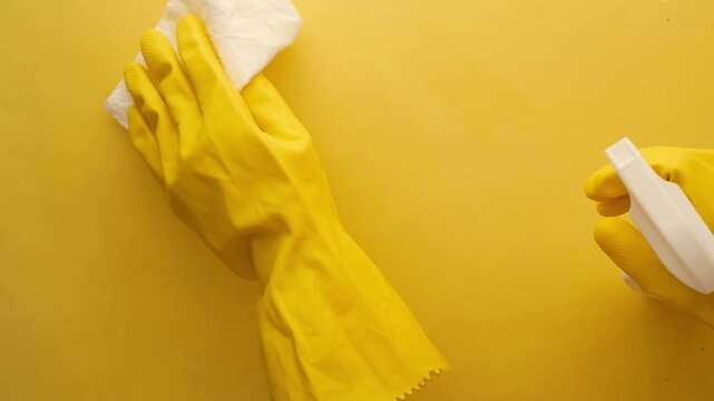 Hands wearing yellow rubber gloves cleaning yellow surface with white cloth and spray bottle