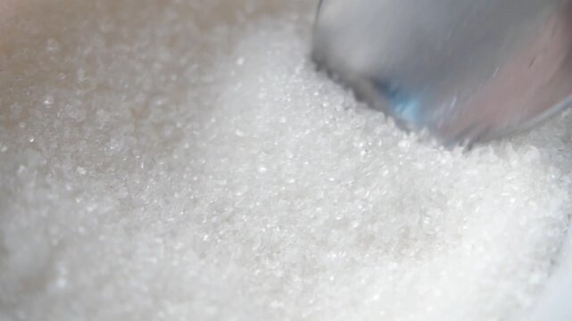 Close up slow motion video of spoonful of refined sugar crystals being scooped from a white bowl, showing its texture and sparkle