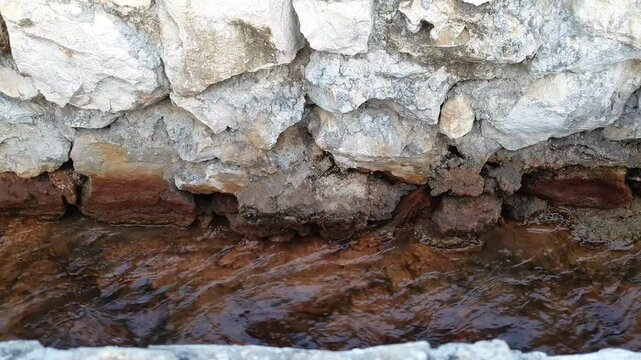 Clear water flowing through a narrow stone drain channel, showing detailed stone wall texture and reddish-brown sediment beneath the water.
