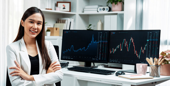 Profile of young Asian businesswoman smiling on happy face with white suit sitting chair against dynamic stock exchange market investing graph on pc showing screen on desk at modern office. Stratagem.