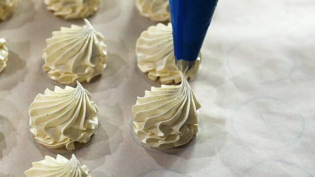 Baker uses a blue piping bag to form meringue shapes on parchment paper, highlighting the detailed pastry preparation in a bright kitchen setting