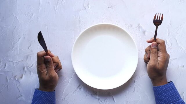 Top-down view of hungry person holding knife and fork on either side of an empty white plate, anticipating a meal, in a static close-up shot of hands and plate on a white table