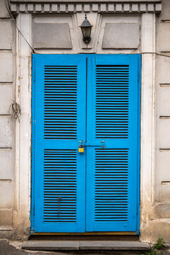 Bright blue wooden shutter door with ventilation slats and metal padlock fills weathered building entrance, framed by aged pale walls and old exterior lantern above closed doorway in urban setting.