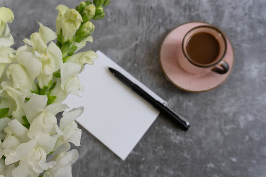 White flowers bloom beside a blank notepad. Black pen rests near a pink saucer. Cup holds warm coffee on a cool gray surface. Stillness suggests creativity or quiet reflection. Perfect for journals