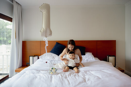 Asian mother and toddler son celebrating first birthday on white bed with cake and balloon