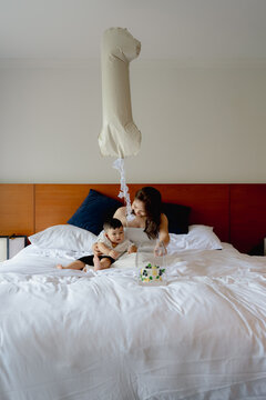 Asian mother and toddler son celebrating first birthday on white bed with cake and balloon