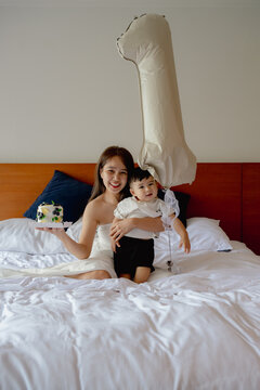 Asian mother and toddler son celebrating first birthday on white bed with cake and balloon