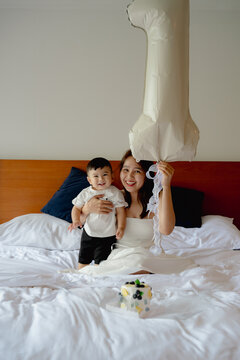 Asian mother and toddler son celebrating first birthday on white bed with cake and balloon