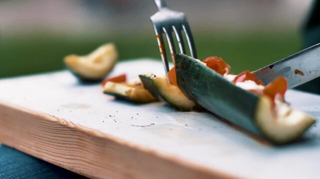 Chef cutting roasted stuffed zucchini on wooden board during outdoor meal preparation.