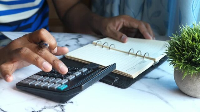 Close up of person using black calculator and flipping pages in daily planner on white marble surface