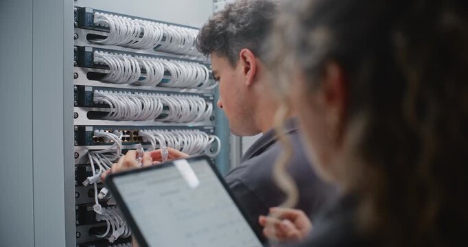 Male Network Engineer Repatches RJ45 Connections While Female Colleague Verifies Work on Tablet, Coordinating Live Telemetry Checks and Structured Cabling Maintenance Inside Modern Data Center.