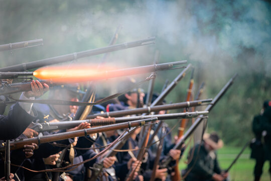 Civil War reenactors portraying Union infantry fire a coordinated volley with rifle muskets, with a bright muzzle flash visible from one weapon and dense black-powder smoke drifting across the firing 