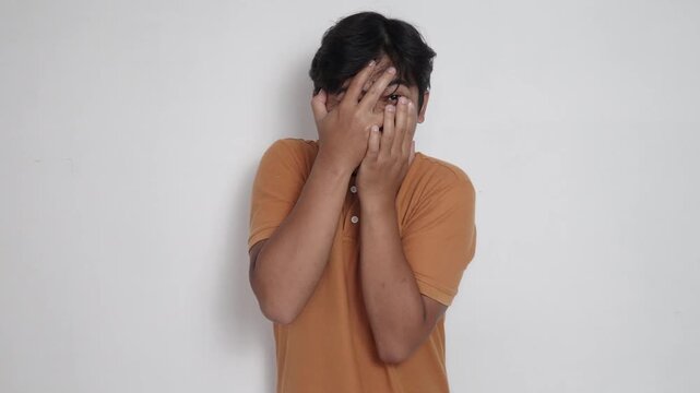 Scared Asian man in orange shirt covering his face with hands and peeking through fingers in fear, frightened, horror reaction, and phobia concept against white background.