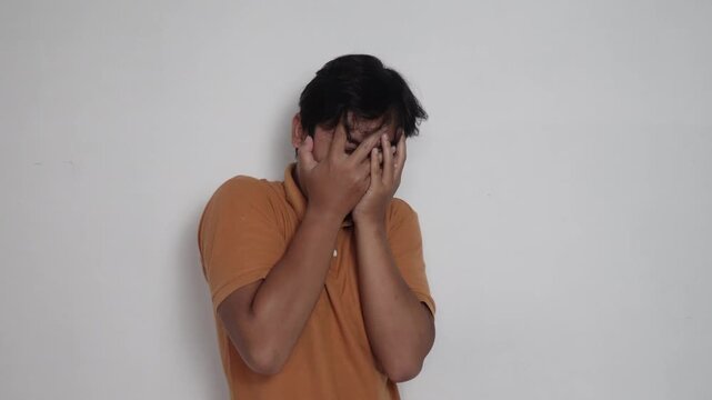 Scared Asian man in orange shirt covering his face with hands and peeking through fingers in fear, frightened, horror reaction, and phobia concept against white background.