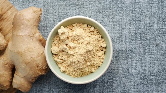 Powdered ginger in small bowl next to fresh ginger root on textured background, slow motion zoom out