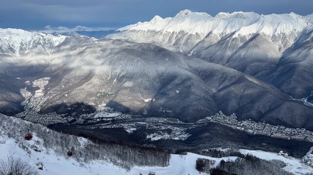Snow-covered mountains with a valley below. The landscape features a mix of rocky peaks and forested areas. A clear sky enhances the winter scenery.