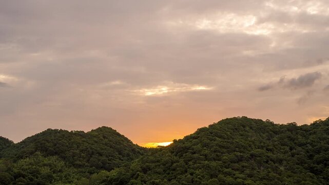 Time lapse view of a beautiful golden sunrise behind a lush green mountain range. Vibrant cloudy sky with orange and yellow morning sunlight over the forest landscape.