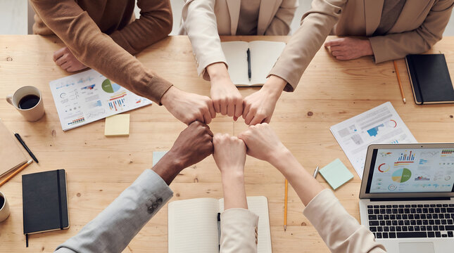 Diverse business team joining fists together in unity and mutual support during a collaborative strategy meeting around a wooden office desk filled with financial documents