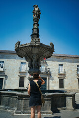 Woman stands before ornate fountain, admiring its sculpted details. Stone structure rises tall, crowned by intricate carvings. Clear blue sky frames historic building behind the fountain © santirj 