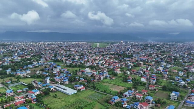 Wide drone perspective of a dense metropolitan valley demonstrating the rural to urban fringe with green fields bordering crowded residential neighborhoods under heavy clouds