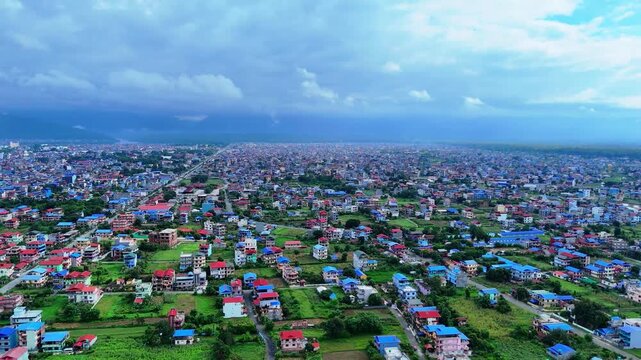 Aerial view over a sprawling South Asian city mixing colorful residential housing with green agricultural plots against a mountain backdrop under an overcast sky