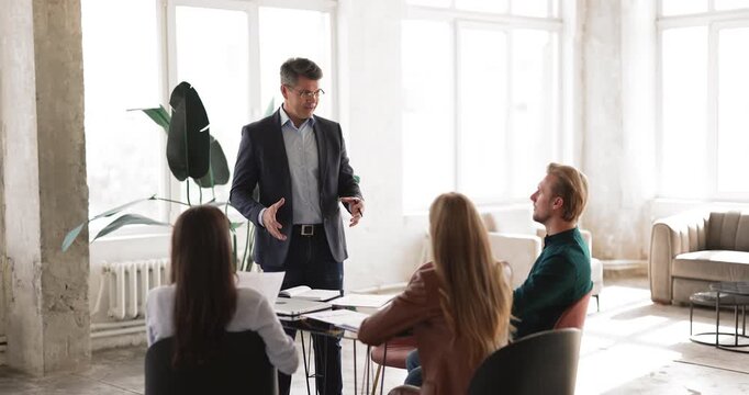 Middle aged male team leader or ceo explaining business strategy presenting development plan to diverse young executives teaching staff on corporate training leading collaborative office conversation