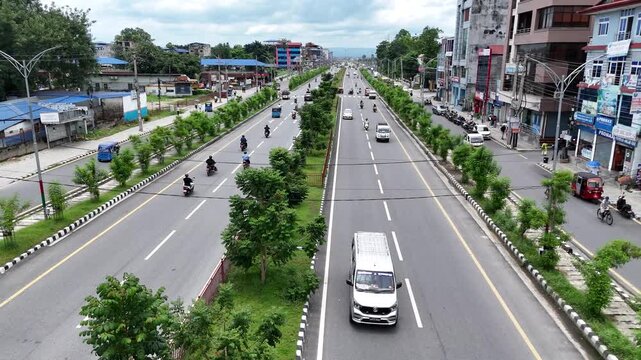 Elevated drone perspective looking down a busy multi-lane divided highway with planted green medians passing commercial storefronts in a Nepalese city