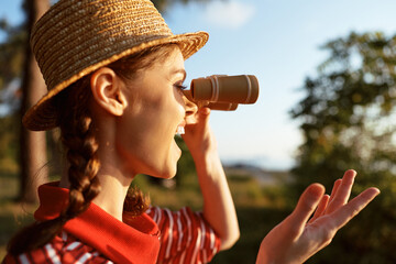 Obraz premium A happy young woman in a straw hat holding binoculars exploring nature lifestyle. She enjoys sunny weather and outdoor activity during a summer trip for travel and adventure.