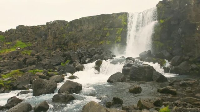 wide angle view of oxararfoss waterfall falling into a rocky riverbed surrounded by green mossy cliffs in thingvellir national park iceland on an overcast day.