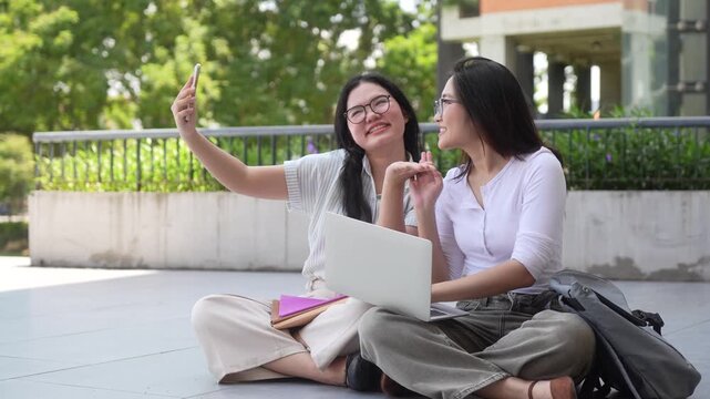Young asian female university students taking selfie with smartphone outdoor on campus, smiling college friends with laptop sitting on ground and making self portrait photo together in university park