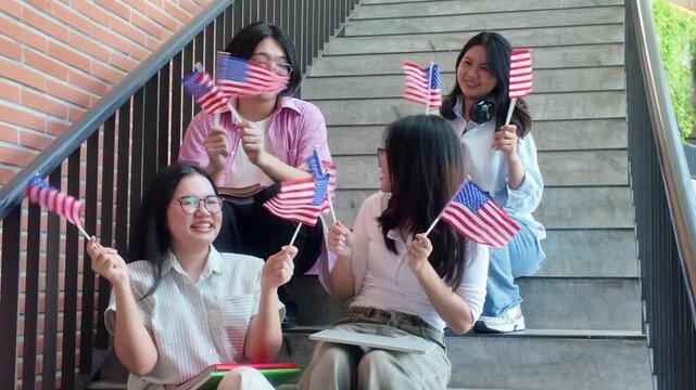 Group of happy asian female university students holding american flags sitting on stairs, smiling college friends celebrating with united states flags at campus building	