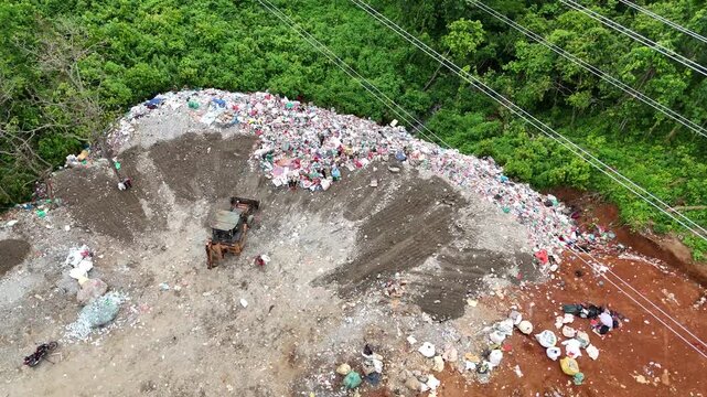 Aerial view of a bulldozer pushing a massive pile of accumulated garbage towards the edge of a green forest at a landfill