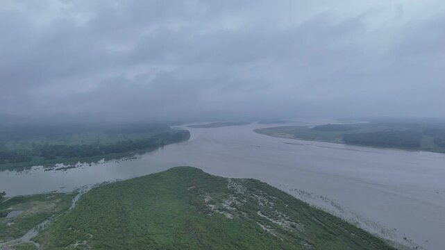 Expansive high altitude aerial view of a wide muddy river bending through a vast green forested floodplain under dark rain clouds