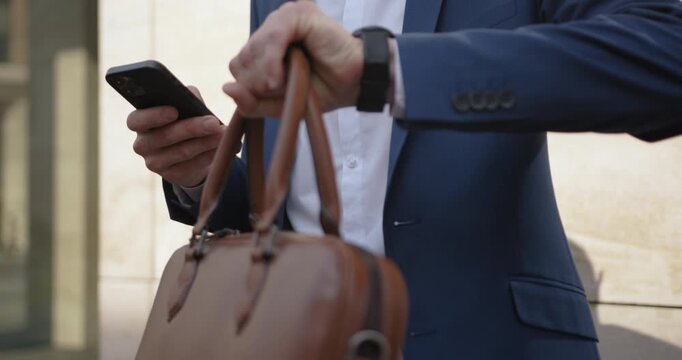 Low angle camera moves upward along a businessman in a blue suit holding a leather briefcase and smartphone outside a modern office building.