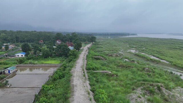 Drone view tracking along a raised dirt levee road separating a rural village from a vast green river floodplain in Nepal
