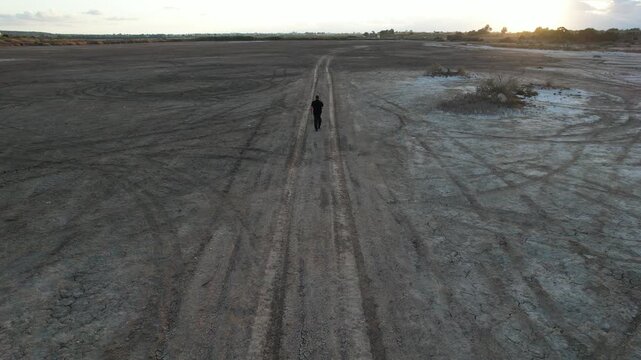 Drone aerial footage of lone man in black walking along a straight dirt path crossing a wide, cracked, arid desert plain lake at sunset. Environmental pollution, dry landscape