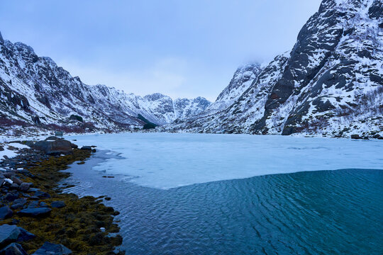 Stunning winter landscape of a partially frozen fjord lake surrounded by snow-covered rugged mountains in Lofoten, Northern Norway. Scenic Arctic mountain valley scenery during a Scandinavian road tri