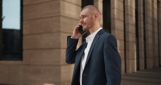 A man in a suit checks his smartphone, raises it to his ear and begins a phone conversation while standing outside a modern building.