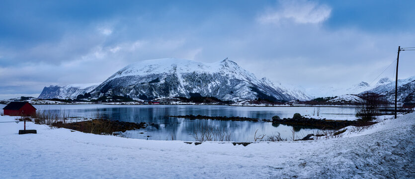 Panoramic winter landscape of Lofoten Islands in Norway. Majestic snow covered mountain reflecting in calm fjord water with traditional red wooden cabin. Scenic Arctic nature panorama of Scandinavian 